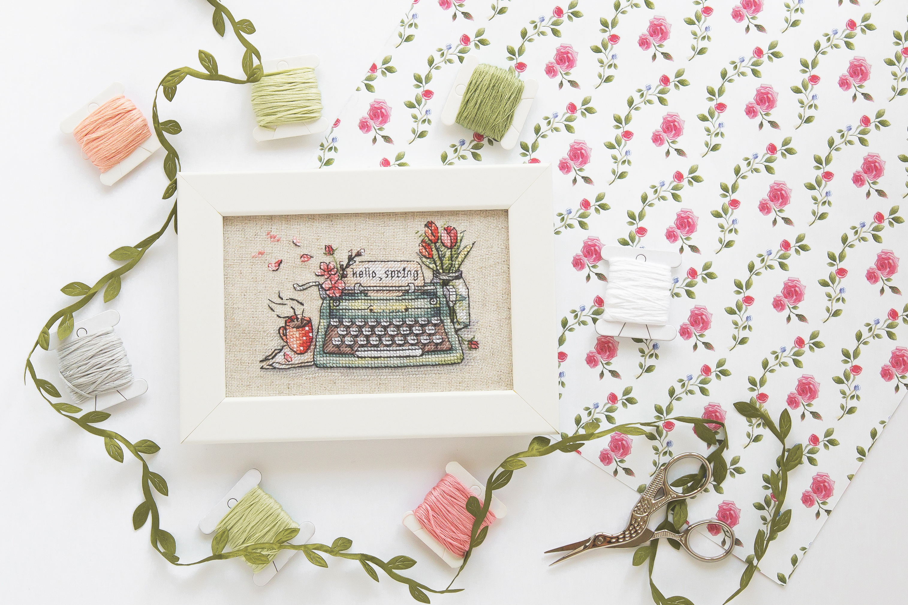 Oprawiony haft krzyżykowy w białej ramce przedstawiający wiosenny motyw, otoczony kolorową muliną i nożyczkami hafciarskimi